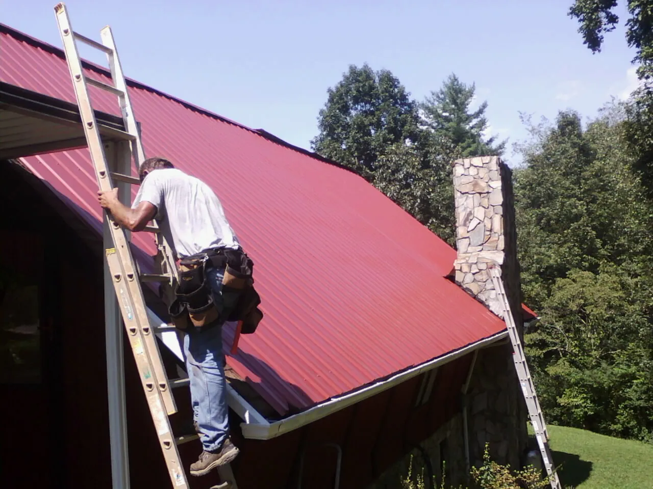 Roofer on a ladder beside a red metal roof and stone chimney — Soffit & Fascia Repair by Roofing Brothers in Wyandanch